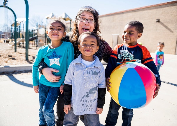 Day care caretaker and kids playing and laughing with ball in school playground. 